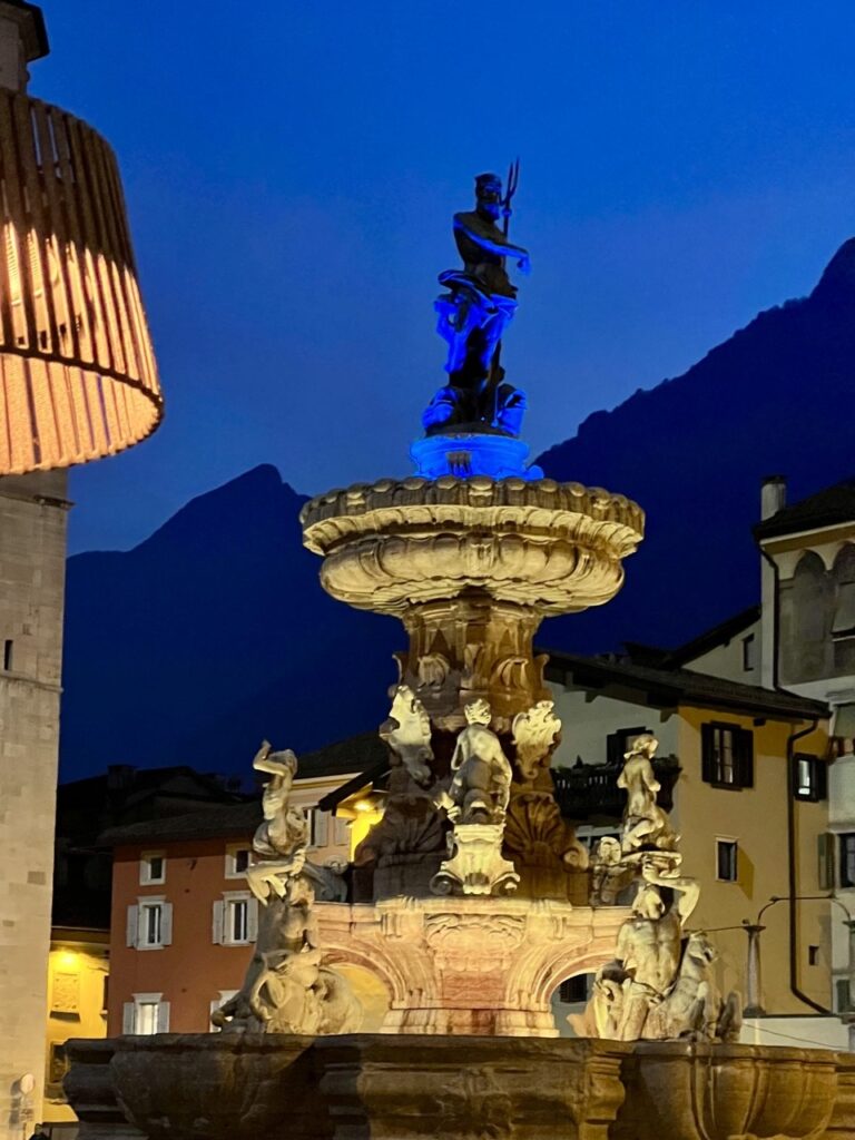The Neptune Fountain in Trento’s Piazza Duomo illuminated at dusk, with the cathedral and mountains framing the historic centre.